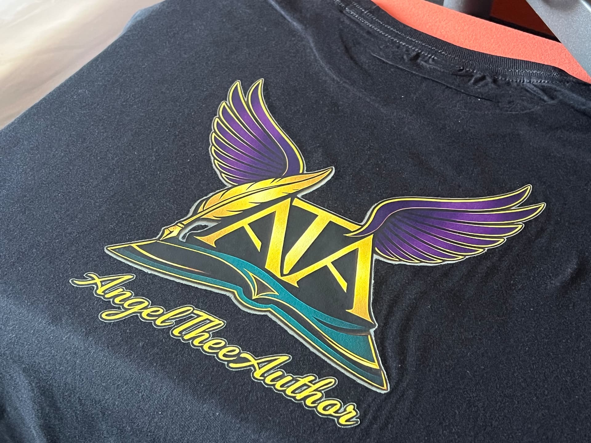 Black shirt pressed with a purple and gold winged book logo for Angel Thee Author